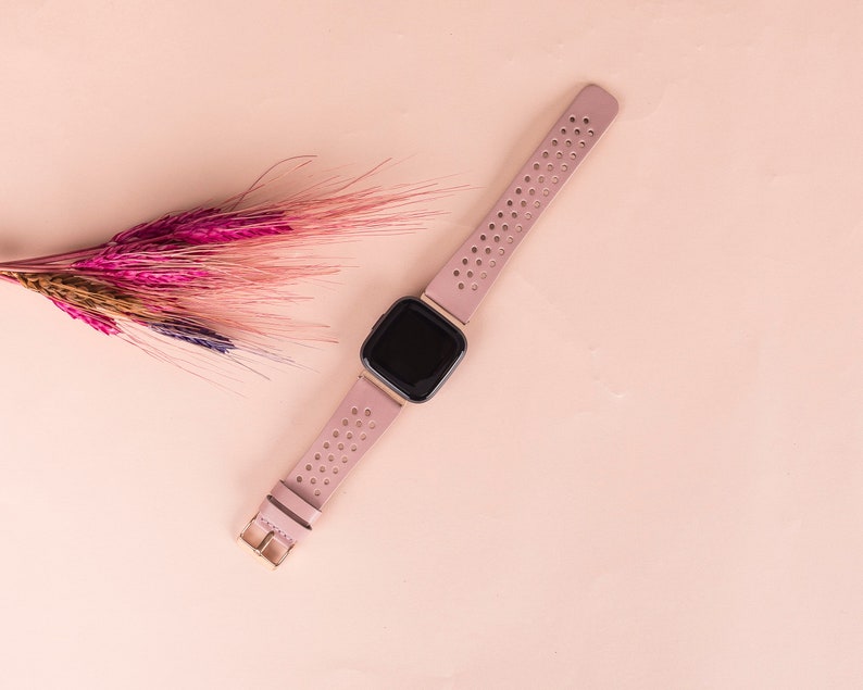 Puede incluir: Un rastreador de actividad f&iacute;sica rosa con una pantalla negra y una banda rosa perforada. El rastreador est&aacute; sobre una superficie rosa claro.