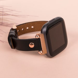 May include: A smartwatch with a black square display and a dark gray casing. The watch features a black leather strap secured with rose gold-toned metal hardware, including a circular stud and connector pieces that attach the strap to the watch body.
