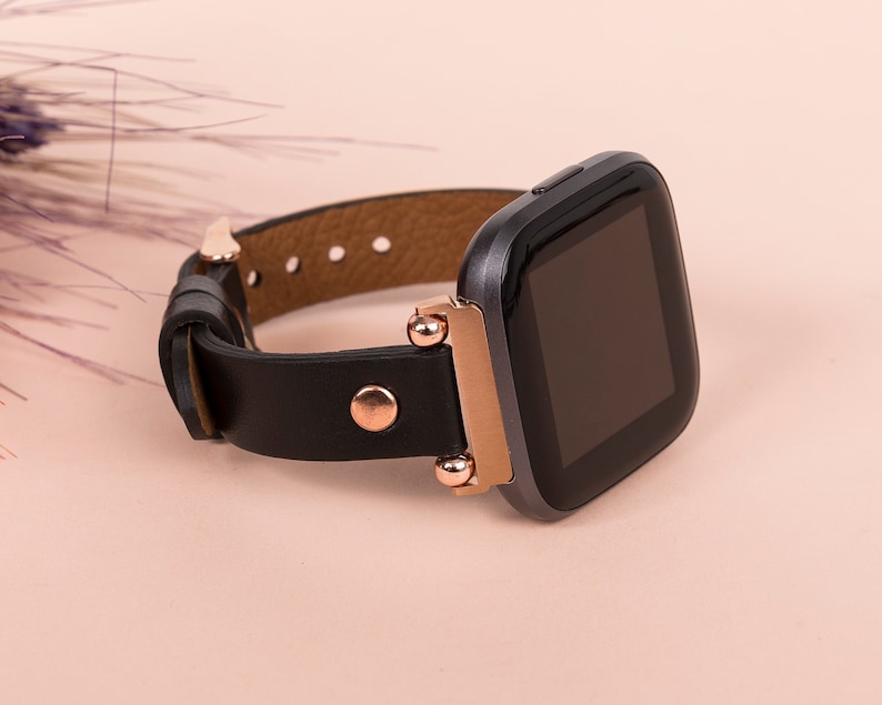 May include: A smartwatch with a black square display and a dark gray casing. The watch features a black leather strap secured with rose gold-toned metal hardware, including a circular stud and connector pieces that attach the strap to the watch body.