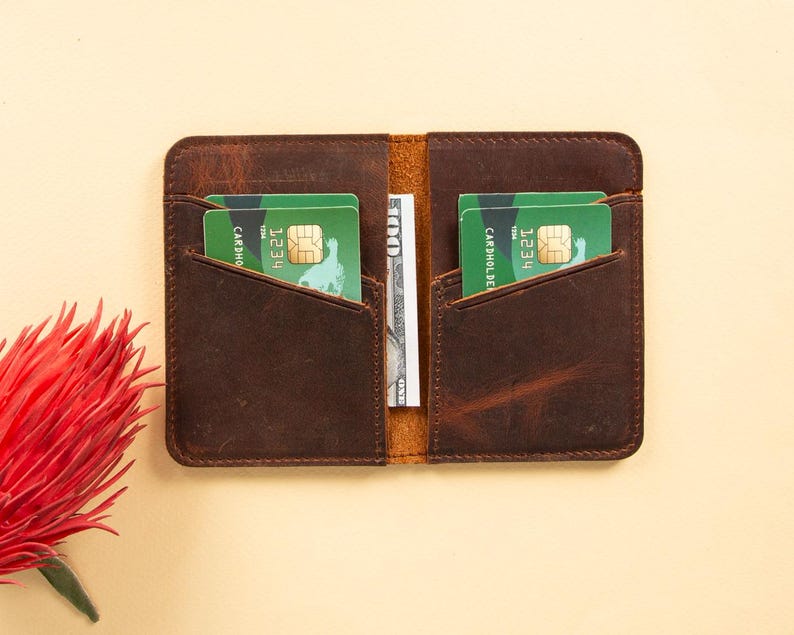 Peut inclure: Un portefeuille en cuir marron avec deux emplacements pour cartes et une poche pour l'argent. Le portefeuille contient deux cartes de cr&eacute;dit vertes avec le texte "CARDHOLDER" et "1234" visible.
