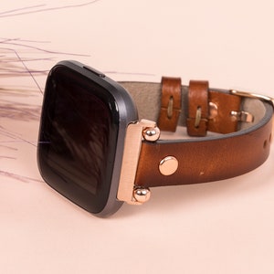 Puede incluir: Una correa de reloj de cuero marr&oacute;n con detalles en oro rosa y una esfera de reloj inteligente negra.