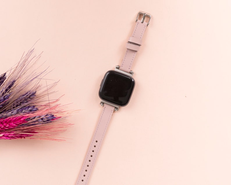May include: A pink leather watch band with a black square watch face. The watch is on a pink background.