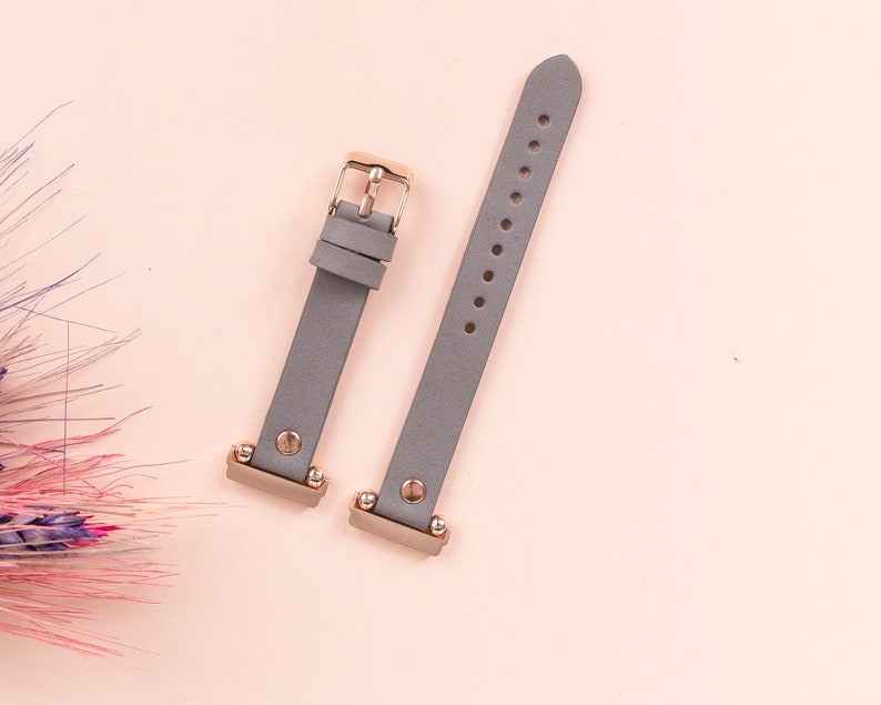 May include: Two gray leather watch bands with rose gold hardware. The bands are flat and have a smooth finish. The hardware includes a buckle and two small studs.