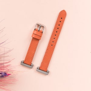 May include: Two orange leather watch straps with silver buckles. The straps are laid out flat on a pink surface.