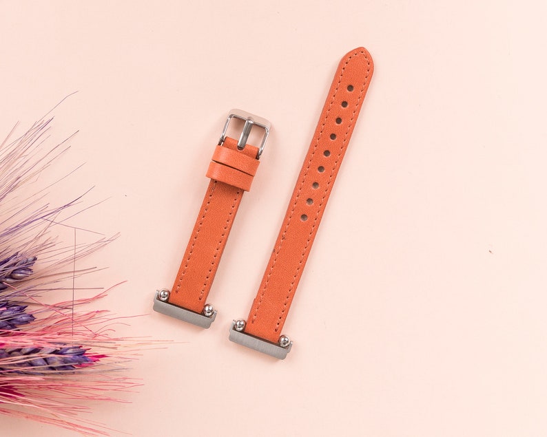 May include: Two orange leather watch straps with silver buckles. The straps are laid out flat on a pink surface.