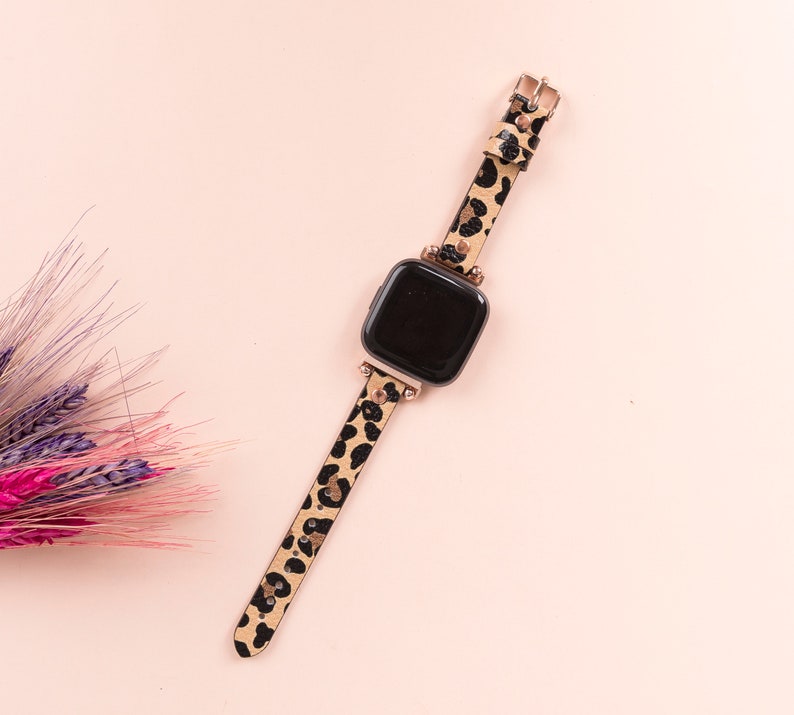 May include: A black and brown leopard print watch band with a black watch face. The band has gold accents and is on a pink background.