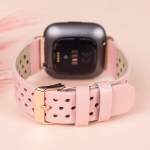 Puede incluir: Una correa de reloj de cuero rosa con herrajes dorados y un dise&ntilde;o perforado. La correa es compatible con los relojes inteligentes Fitbit Versa.
