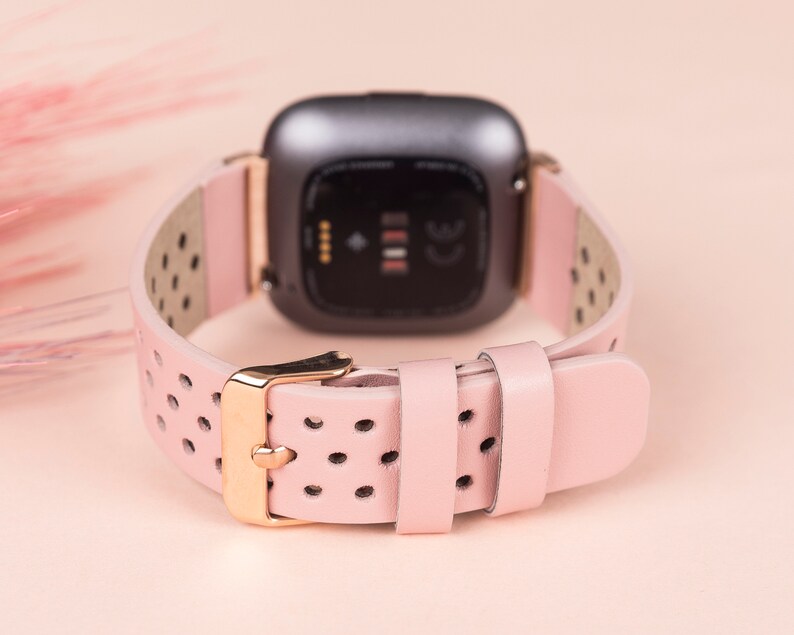 Puede incluir: Una correa de reloj de cuero rosa con herrajes dorados y un dise&ntilde;o perforado. La correa es compatible con los relojes inteligentes Fitbit Versa.