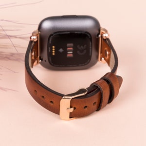Puede incluir: Una correa de reloj de cuero marr&oacute;n con herrajes de oro rosa y un reloj inteligente negro con una pantalla negra. La correa del reloj tiene un cierre de hebilla.