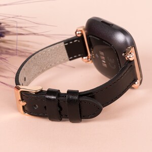 May include: Black leather watch strap with rose gold hardware and a buckle fastening. The strap is compatible with Fitbit Versa smartwatches.