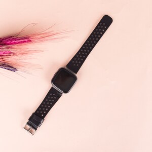 May include: A black fitness tracker with a perforated black band on a pink background.