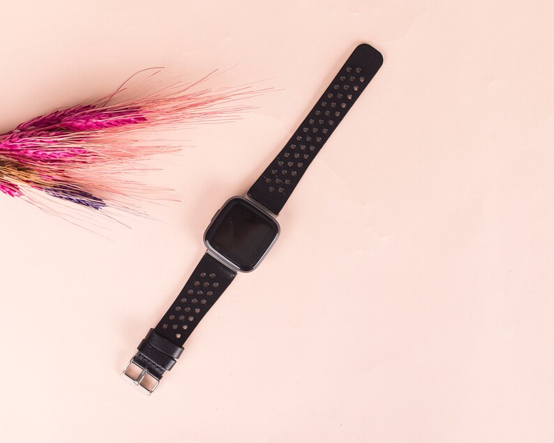 May include: A black fitness tracker with a perforated black band on a pink background.