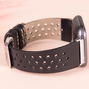 May include: Black leather watch strap with a perforated design and a silver buckle. The strap is shown against a light pink background.