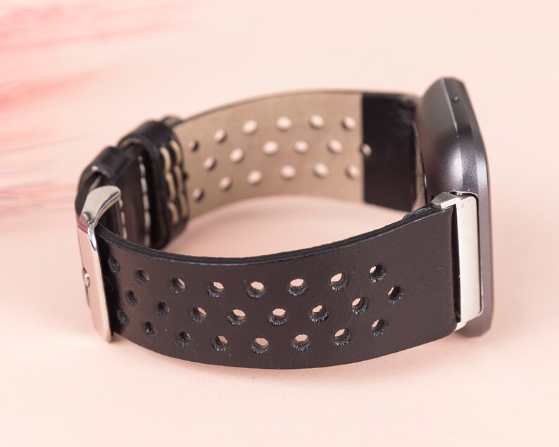 May include: Black leather watch strap with a perforated design and a silver buckle. The strap is shown against a light pink background.