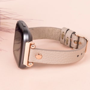 May include: A gray smartwatch with a light gray leather band and rose gold accents. The band has a buckle closure.