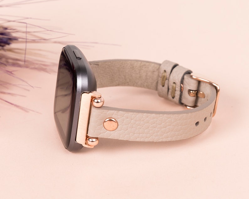 May include: A gray smartwatch with a light gray leather band and rose gold accents. The band has a buckle closure.