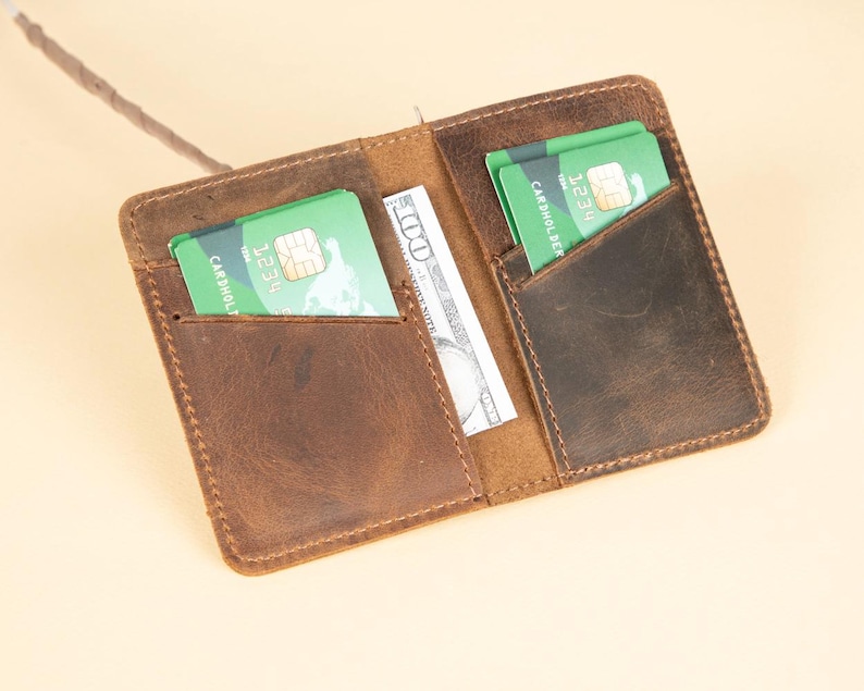 Peut inclure: Un portefeuille en cuir marron avec deux emplacements pour cartes et un porte-billets. Le portefeuille a un aspect vieilli et est ouvert pour montrer l'int&eacute;rieur.