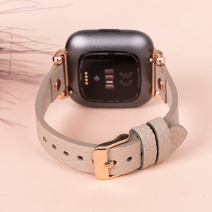 May include: A gray and rose gold smartwatch band with a textured leather strap. The band is designed for a smartwatch with a square face.