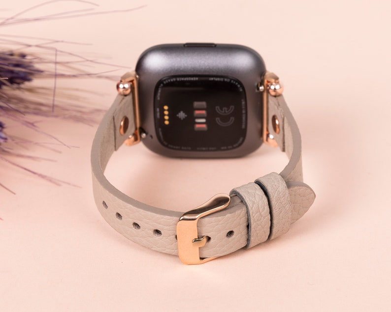 May include: A gray and rose gold smartwatch band with a textured leather strap. The band is designed for a smartwatch with a square face.
