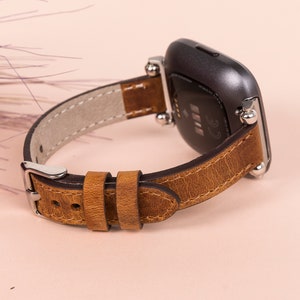 K&ouml;nnte beinhalten: Ein braunes Lederarmband mit silbernen Beschl&auml;gen und einer silbernen Schlie&szlig;e. Das Armband ist f&uuml;r eine quadratische Smartwatch konzipiert.