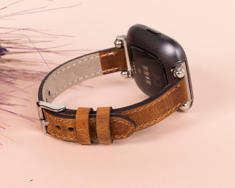 K&ouml;nnte beinhalten: Ein braunes Lederarmband mit silbernen Beschl&auml;gen und einer silbernen Schlie&szlig;e. Das Armband ist f&uuml;r eine quadratische Smartwatch konzipiert.