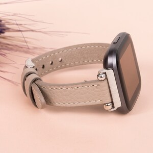 Puede incluir: Una correa de reloj de cuero gris con detalles plateados y una esfera de reloj cuadrada negra.