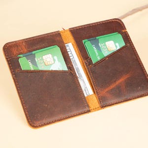 Peut inclure: Un portefeuille en cuir marron avec deux emplacements pour cartes et une poche pour l'argent. Le portefeuille est ouvert et contient deux cartes de cr&eacute;dit vertes &agrave; l'int&eacute;rieur.