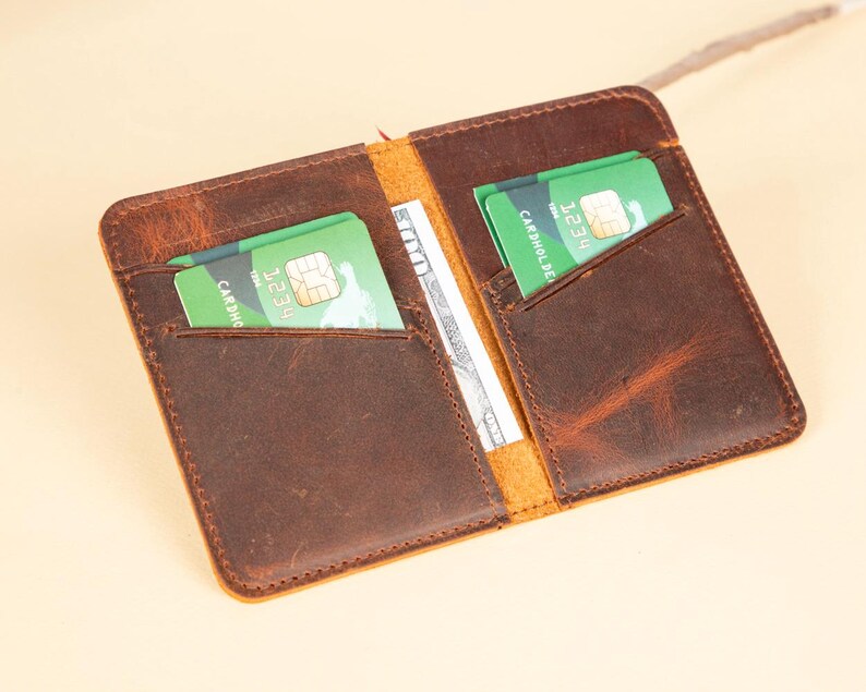 Peut inclure: Un portefeuille en cuir marron avec deux emplacements pour cartes et une poche pour l'argent. Le portefeuille est ouvert et contient deux cartes de cr&eacute;dit vertes &agrave; l'int&eacute;rieur.