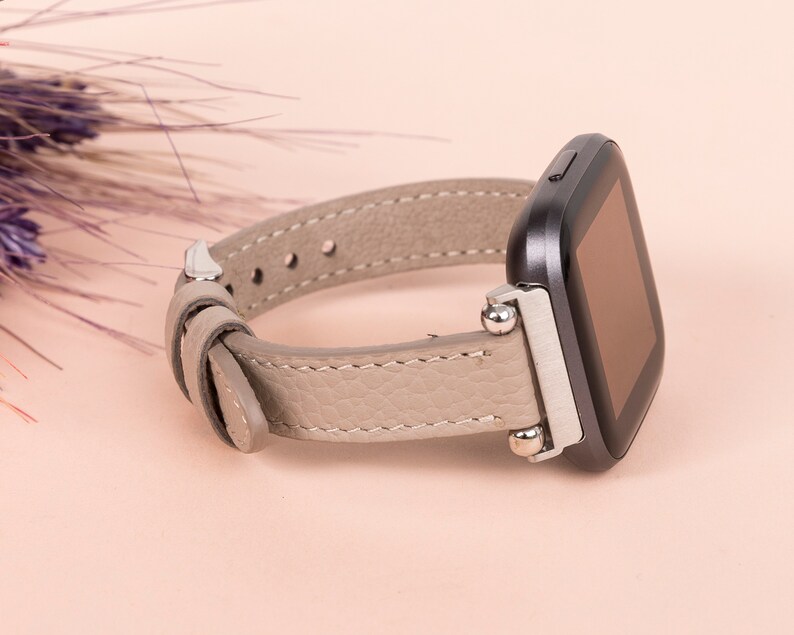 Puede incluir: Una correa de reloj de cuero gris con detalles plateados y un reloj inteligente cuadrado negro.