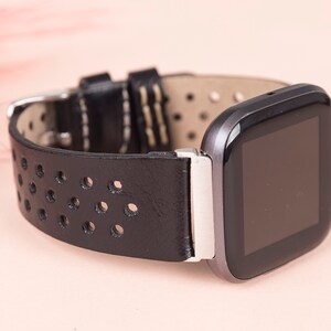 May include: Black leather watch strap with a perforated design and a silver buckle. The strap is attached to a black and silver smartwatch with a square face.