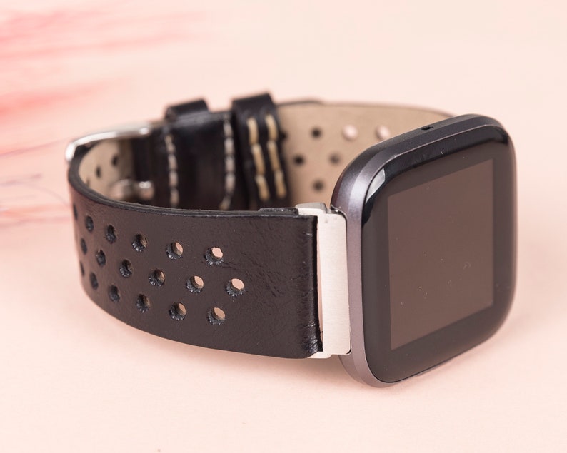 May include: Black leather watch strap with a perforated design and a silver buckle. The strap is attached to a black and silver smartwatch with a square face.