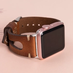 May include: A rose gold Apple Watch with a brown leather band. The band has a unique design with a cut-out section and stitching.