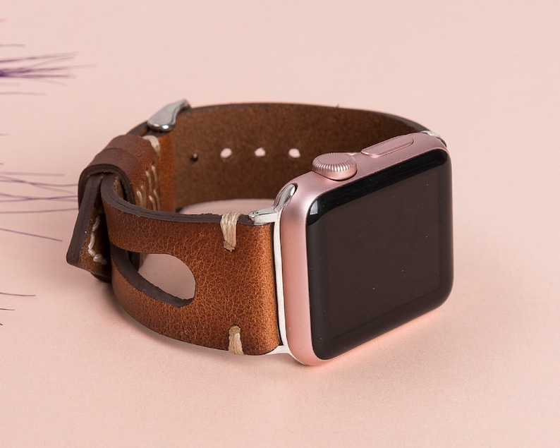 May include: A rose gold Apple Watch with a brown leather band. The band has a unique design with a cut-out section and stitching.
