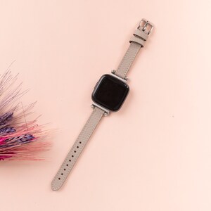 Puede incluir: Una correa de reloj de cuero gris con una hebilla plateada y una esfera de reloj cuadrada negra.