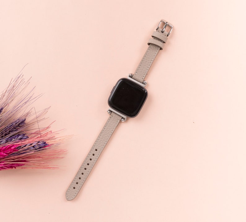 Puede incluir: Una correa de reloj de cuero gris con una hebilla plateada y una esfera de reloj cuadrada negra.