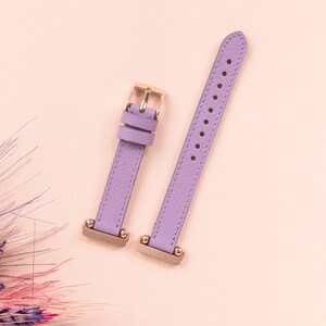 May include: Two purple leather watch bands with gold hardware. The bands are on a light pink background.