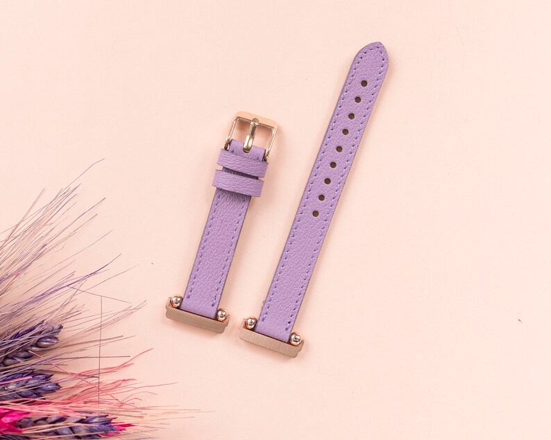 May include: Two purple leather watch bands with gold hardware. The bands are on a light pink background.