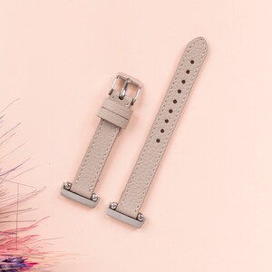 Puede incluir: Dos correas de reloj de cuero gris claro con hebillas plateadas. Las correas est&aacute;n sobre un fondo rosa claro.