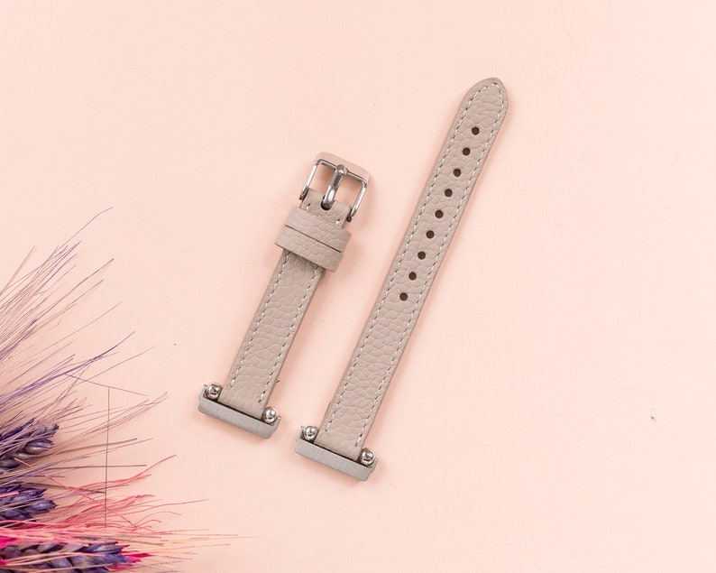Puede incluir: Dos correas de reloj de cuero gris claro con hebillas plateadas. Las correas est&aacute;n sobre un fondo rosa claro.