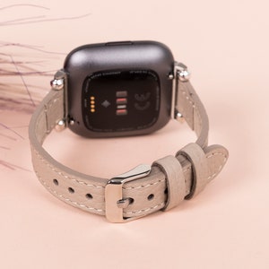 Puede incluir: Un reloj inteligente gris con una correa de cuero gris claro. La esfera del reloj es negra y tiene un peque&ntilde;o punto blanco en el centro.