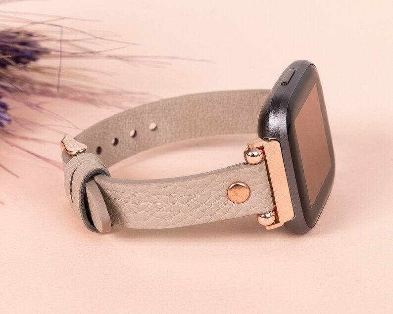 May include: A gray leather watch band with rose gold accents and a black square watch face.