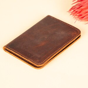 Peut inclure: Un portefeuille en cuir marron avec une finition vieillie. Le portefeuille est ferm&eacute; et pos&eacute; &agrave; plat sur une surface beige clair.