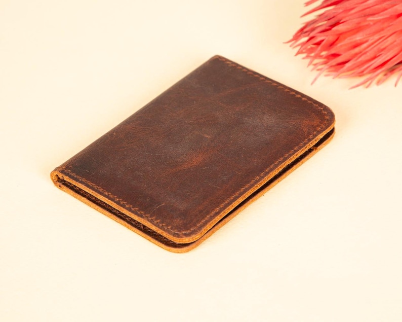 Peut inclure: Un portefeuille en cuir marron avec une finition vieillie. Le portefeuille est ferm&eacute; et pos&eacute; &agrave; plat sur une surface beige clair.