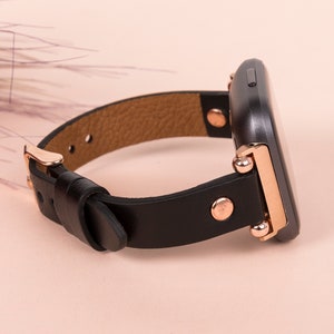 May include: A smartwatch with a black rectangular face and a black leather strap. The watch features rose gold metal hardware, including a buckle, side studs, and connectors that attach the strap to the watch body. The interior of the strap is tan.