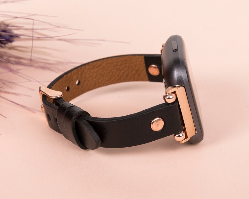 May include: A smartwatch with a black rectangular face and a black leather strap. The watch features rose gold metal hardware, including a buckle, side studs, and connectors that attach the strap to the watch body. The interior of the strap is tan.