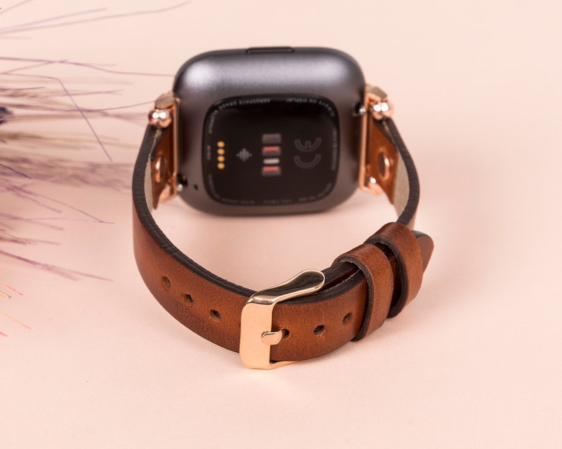 Puede incluir: Una correa de reloj de cuero marr&oacute;n con herrajes de oro rosa y un reloj inteligente negro. La correa del reloj tiene un cierre de hebilla y est&aacute; dise&ntilde;ada para un reloj inteligente de forma cuadrada.