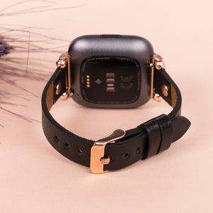 May include: A smartwatch with a dark gray casing and a black leather strap. The watch features rose gold-toned metal hardware, including the buckle and the connectors attaching the strap to the watch body. The back of the watch is visible.
