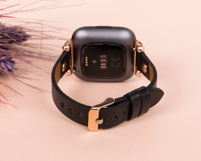 May include: A smartwatch with a dark gray casing and a black leather strap. The watch features rose gold-toned metal hardware, including the buckle and the connectors attaching the strap to the watch body. The back of the watch is visible.