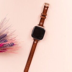 Puede incluir: Una correa de reloj de cuero marr&oacute;n con hebilla dorada y una esfera de reloj cuadrada negra.