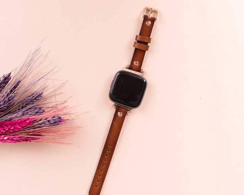 Puede incluir: Una correa de reloj de cuero marr&oacute;n con hebilla dorada y una esfera de reloj cuadrada negra.